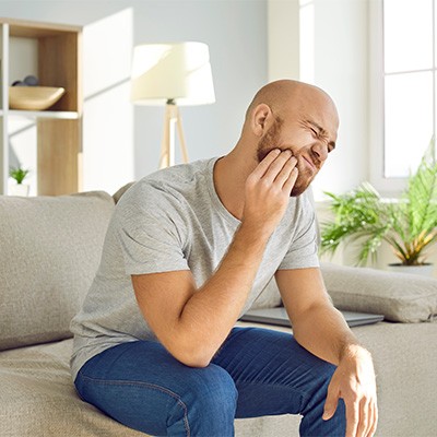 Man on couch experiencing painful jaw holding his cheek